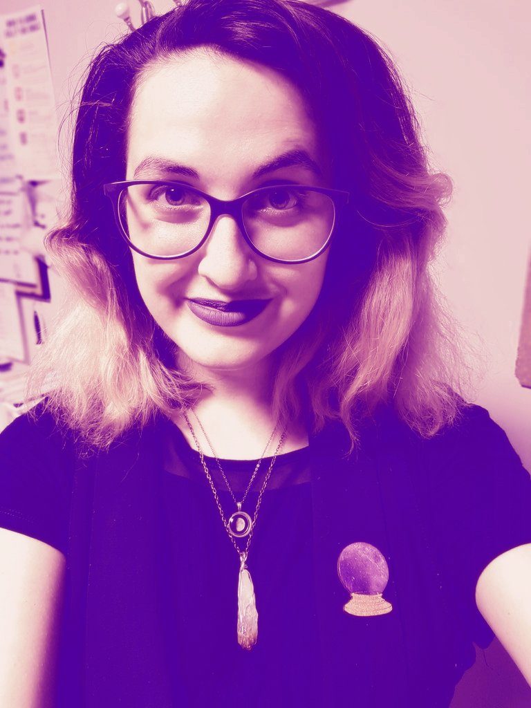 A selfie of Jillian. She has dark to light ombre hair. She is wearing glasses, dark lipstick, 2 necklaces, a crystal ball pin and a black tshirt. The colour has been edited to be only purple.